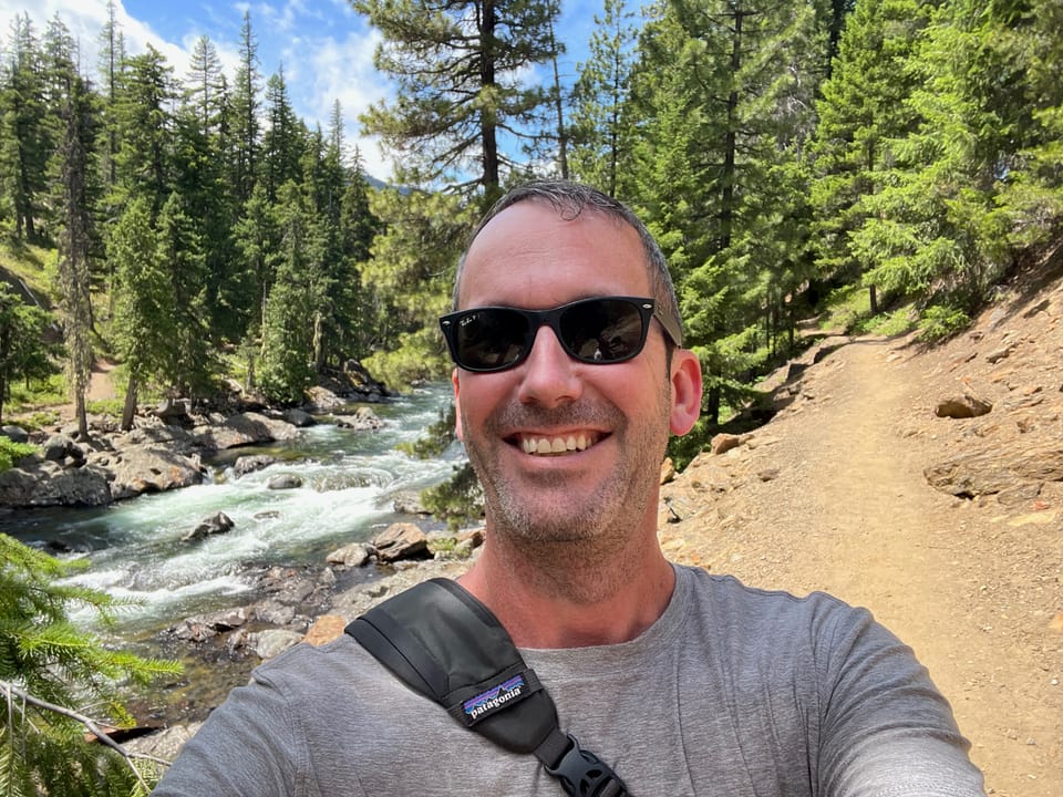 Justin, in sunglasses, smiles for a selfie on a forest trail beside a rocky river, surrounded by tall evergreen trees under a partly cloudy sky.