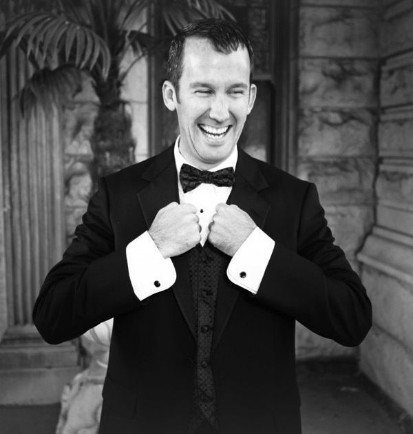 Justin in a tuxedo adjusting his bow tie with a big smile.