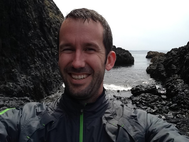 Justin smiling in a gray and black jacket, taking a selfie with dramatic black volcanic rock formations and the ocean behind him on an overcast day