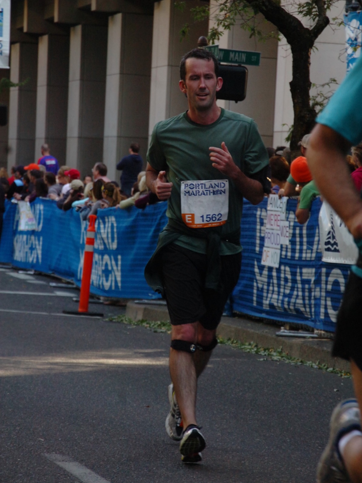 Justin mid-stride in green running gear during the 2012 Portland Marathon, passing cheering crowds lined behind blue barriers on Main Street