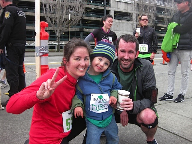 Justin and Audrey kneeling with toddler Wencel between them at a race finish, all smiling. Audrey throws up a peace sign while Justin holds coffee, and Wencel wears his race bib proudly