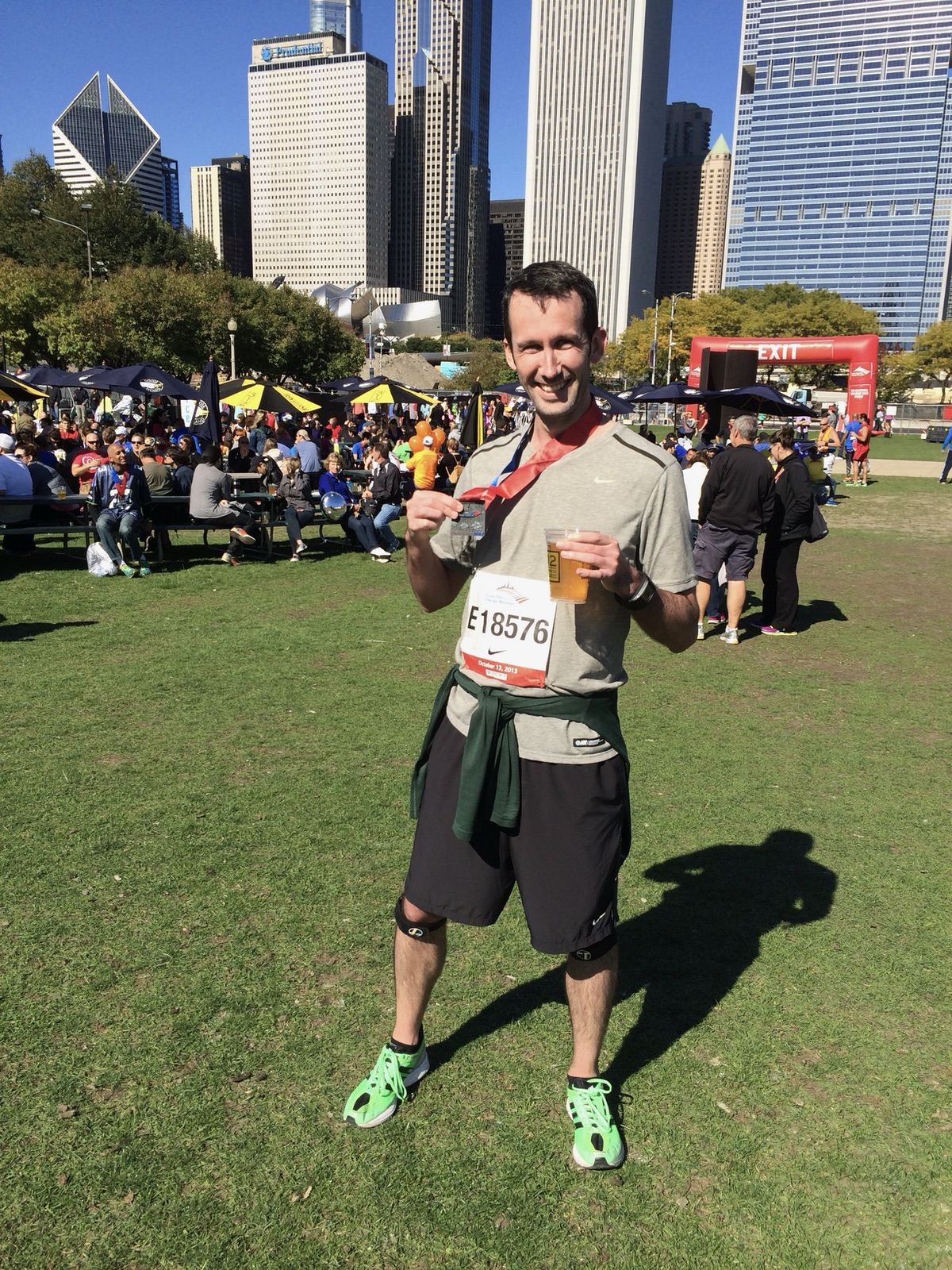 Justin holding his finisher medal and a beer at the Chicago Marathon post-race festival, with the city skyline and crowds behind him on a sunny day