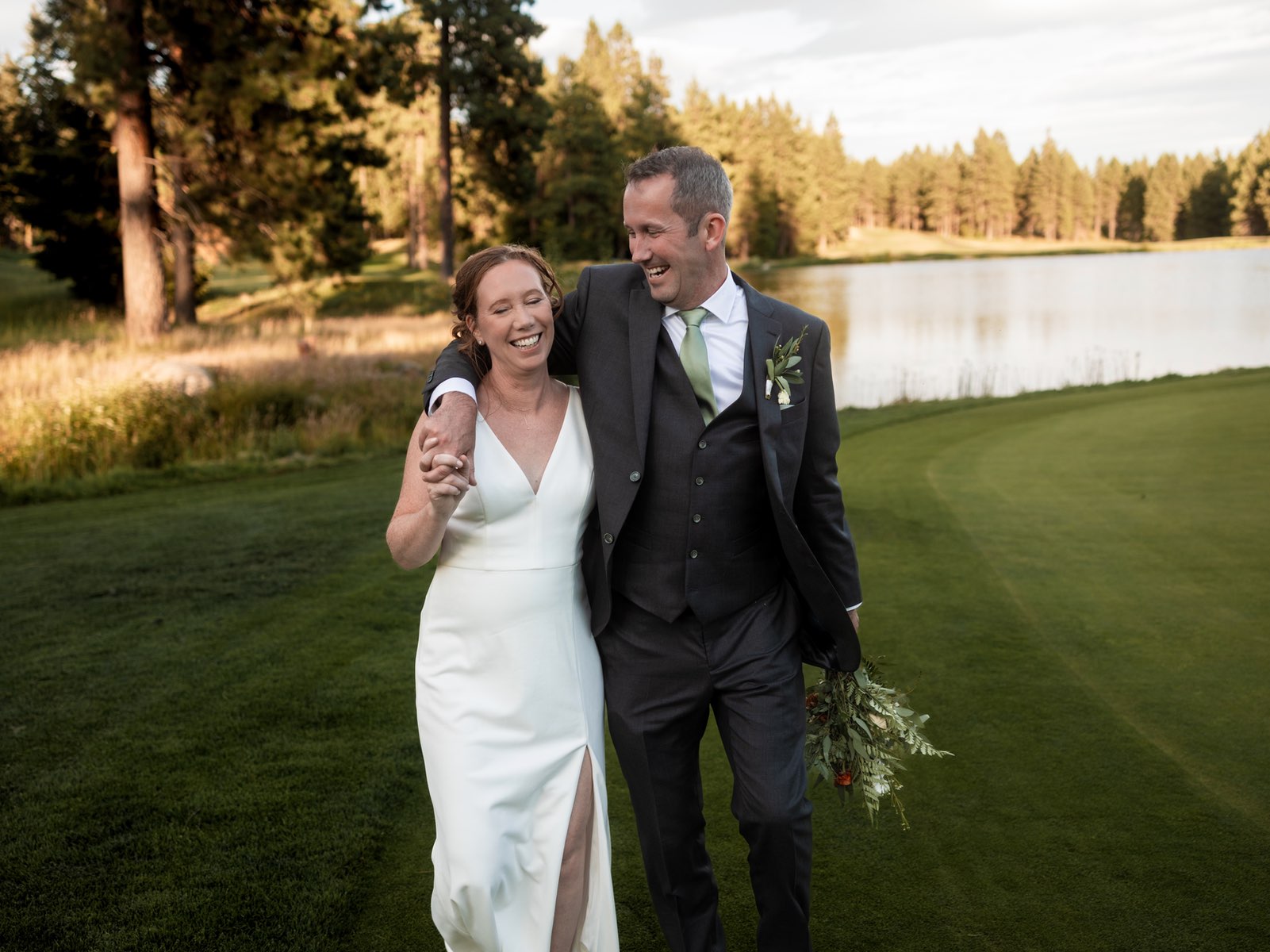 Pam and Justin walk arm-in-arm across a manicured lawn at golden hour, both smiling and laughing together. The bride wears a simple white V-neck gown with a side slit and holds a greenery bouquet, while the groom wears a dark gray suit with vest and light green tie, with a small boutonniere. Behind them is a serene pond surrounded by pine trees with warm afternoon sunlight filtering through.