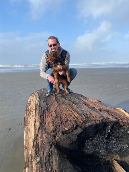 Justin crouching on a large piece of driftwood on a sandy beach. He is holding Olive, a small live and tan English Cocker Spaniel. Justin is wearing sunglasses, a light gray top, and blue jeans. The setting is a wide, open beach with wet sand, and the ocean is visible in the background with small waves breaking on the shore. The sky is mostly blue with some white clouds scattered throughout. The driftwood appears to be a large weathered log or tree trunk washed up on the beach. The scene depicts a clear, pleasant day at the beach, with the Justin and Olive positioned atop the natural wooden landmark.