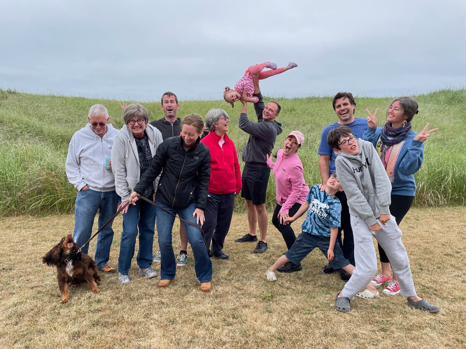 A cheerful group of eleven people posing informally and goofily on a patch of dry grass with tall green dune grasses behind them. The group includes members of Justin and Pam's families. Olive stands with them on the left - barking at the drone taking the photo, while Nate playfully holds a Layla overhead. Despite the overcast sky, the group displays animated, playful poses and expressions, creating a joyful atmosphere during what seems to be a casual outdoor gathering.