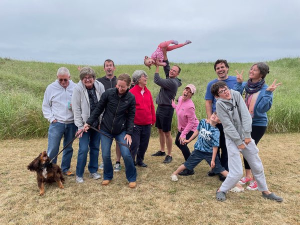 A cheerful group of eleven people posing informally and goofily on a patch of dry grass with tall green dune grasses behind them. The group includes members of Justin and Pam's families. Olive stands with them on the left - barking at the drone taking the photo, while Nate playfully holds a Layla overhead. Despite the overcast sky, the group displays animated, playful poses and expressions, creating a joyful atmosphere during what seems to be a casual outdoor gathering.