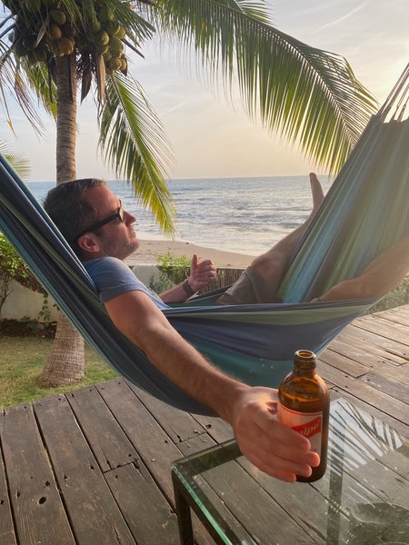 Justin relaxes in a blue hammock on a wooden deck overlooking the beach, holding a bottled beverage. He's wearing sunglasses and casual clothing with their feet up. A coconut palm tree frames the scene to the left, with the ocean visible in the background during what appears to be sunset or early evening. The setting conveys a peaceful tropical vacation atmosphere with golden hour lighting across the shoreline.