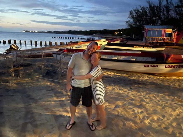 Justin and Pam stand embracing on a sandy beach at dusk, with fishing boats or small watercraft visible behind them on the shore. Justin wears a light-colored shirt with dark shorts, while the Pam wears a white and black striped dress or outfit. The scene is illuminated by twilight with a blue-gray sky, and what appears to be wooden pilings or a dock structure can be seen extending into the water. Soft lighting highlights the sand and boats, creating a warm evening beach atmosphere with some distant lights visible across the water.