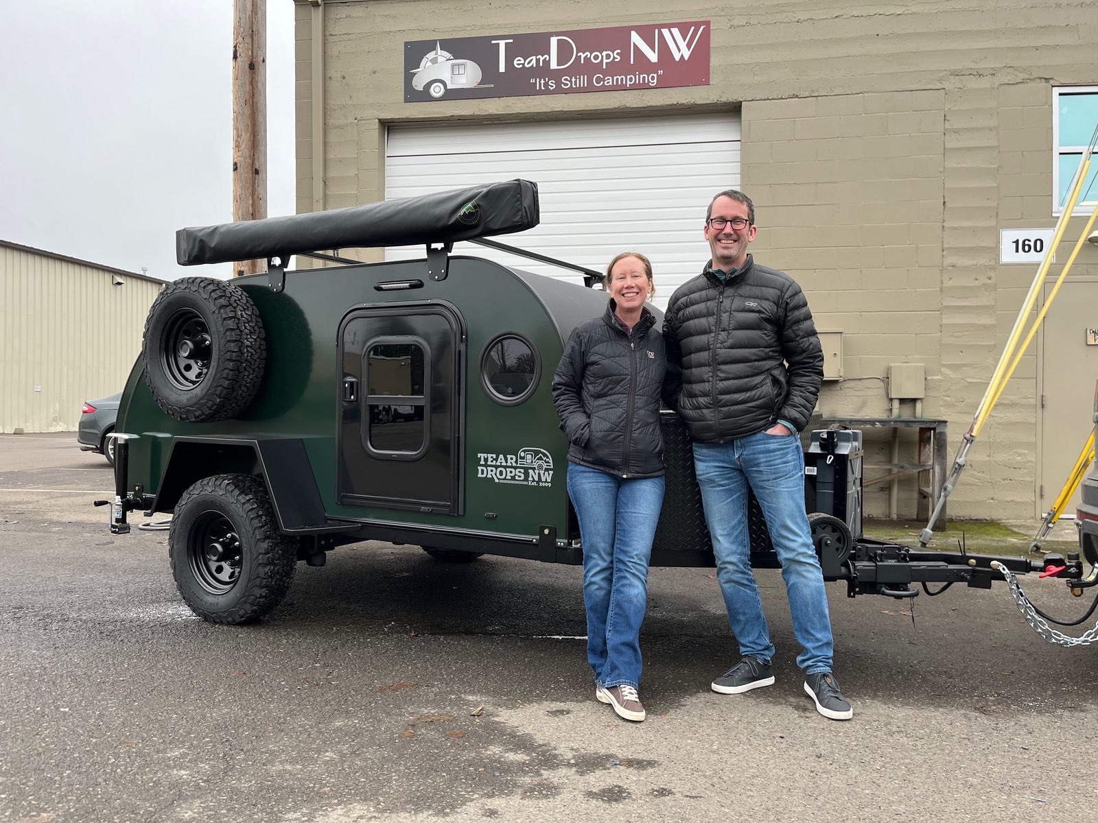 A green teardrop trailer with a rooftop tent parked outside the TearDrops NW facility. Pam and Justin are standing beside the trailer, smiling at the camera. The building behind them has a sign that reads 'TearDrops NW' with the tagline 'It's Still Camping!' The compact trailer features black wheels, a rear hatch, and appears designed for outdoor adventures.