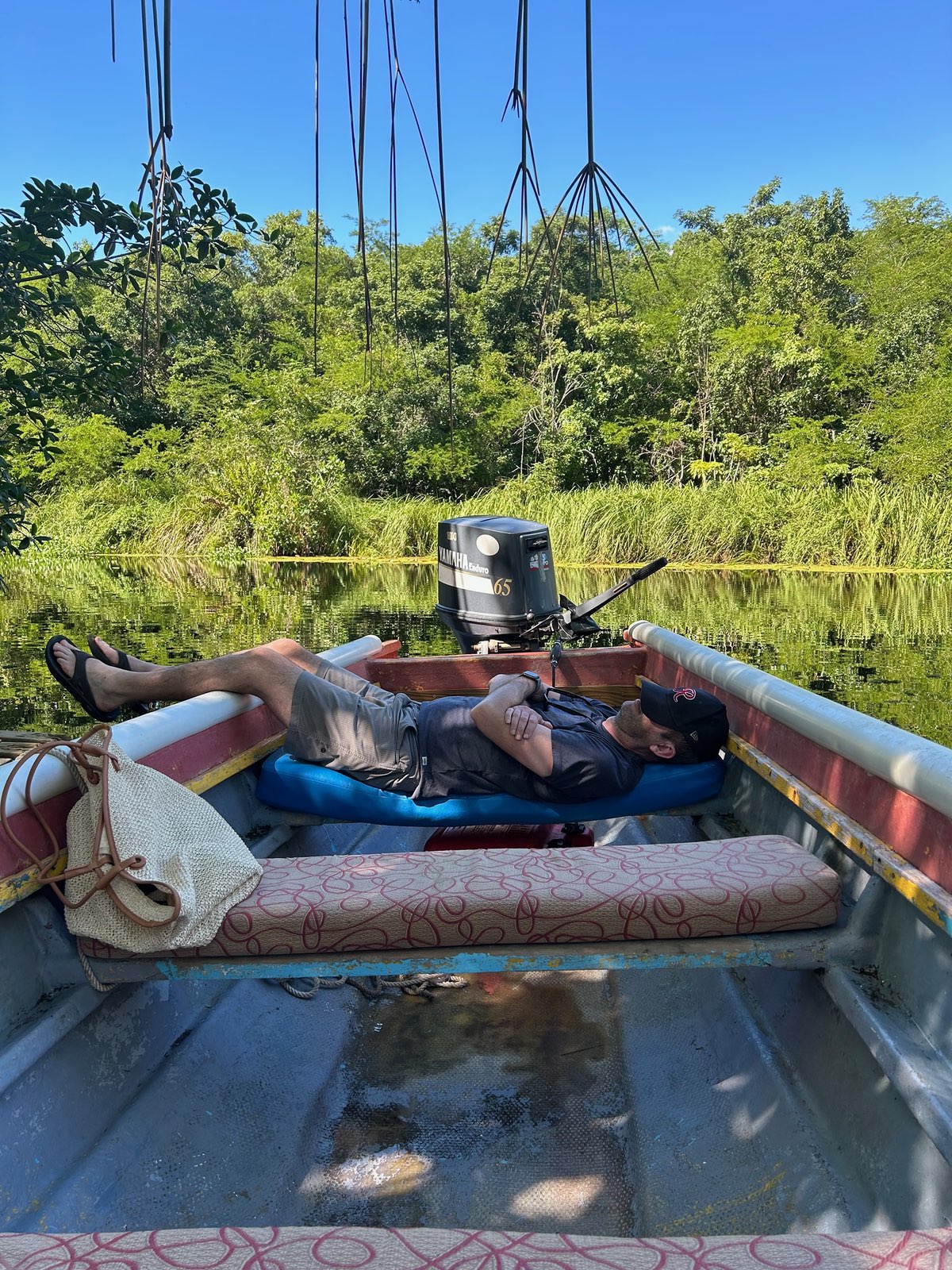 Justin is relaxing in a small fishing boat on calm water on a river in Jamaica. He's lying back on cushioned boat seats with his legs stretched out, appearing to be napping or resting. The boat has an outboard motor and is surrounded by lush green vegetation reflecting in the still water. The scene captures a peaceful moment of leisure.