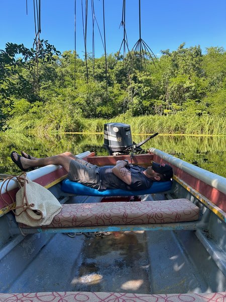 Justin is relaxing in a small fishing boat on calm water on a river in Jamaica. He's lying back on cushioned boat seats with his legs stretched out, appearing to be napping or resting. The boat has an outboard motor and is surrounded by lush green vegetation reflecting in the still water. The scene captures a peaceful moment of leisure.