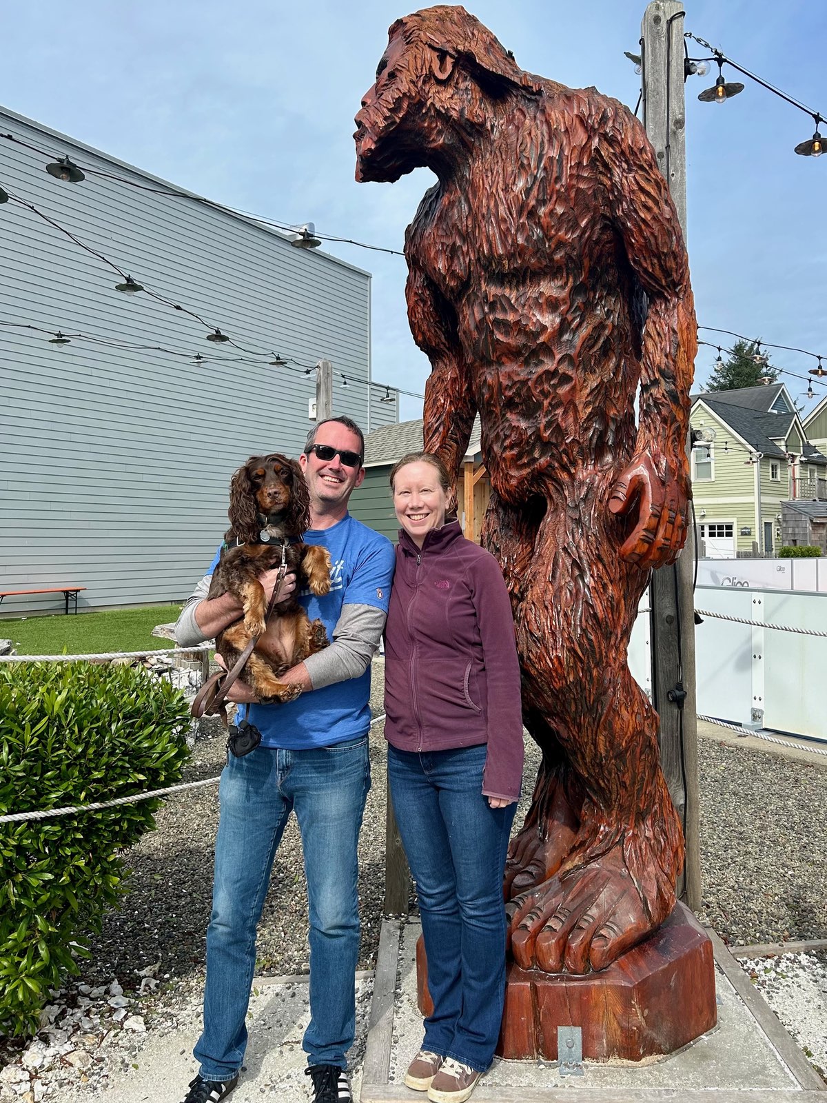 A tall wooden sasquatch/bigfoot sculpture standing next to Justin and Pam. Justin is holding Olive, their brown English Cocker Spaniel. The wooden carving, showing a standing bigfoot figure with detailed fur texture, towers over the visitors.